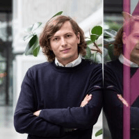 Portrait de Guilhem, consultant en marketing digital et rédaction SEO, bras croisés devant une baie vitrée avec plante en arrière-plan.